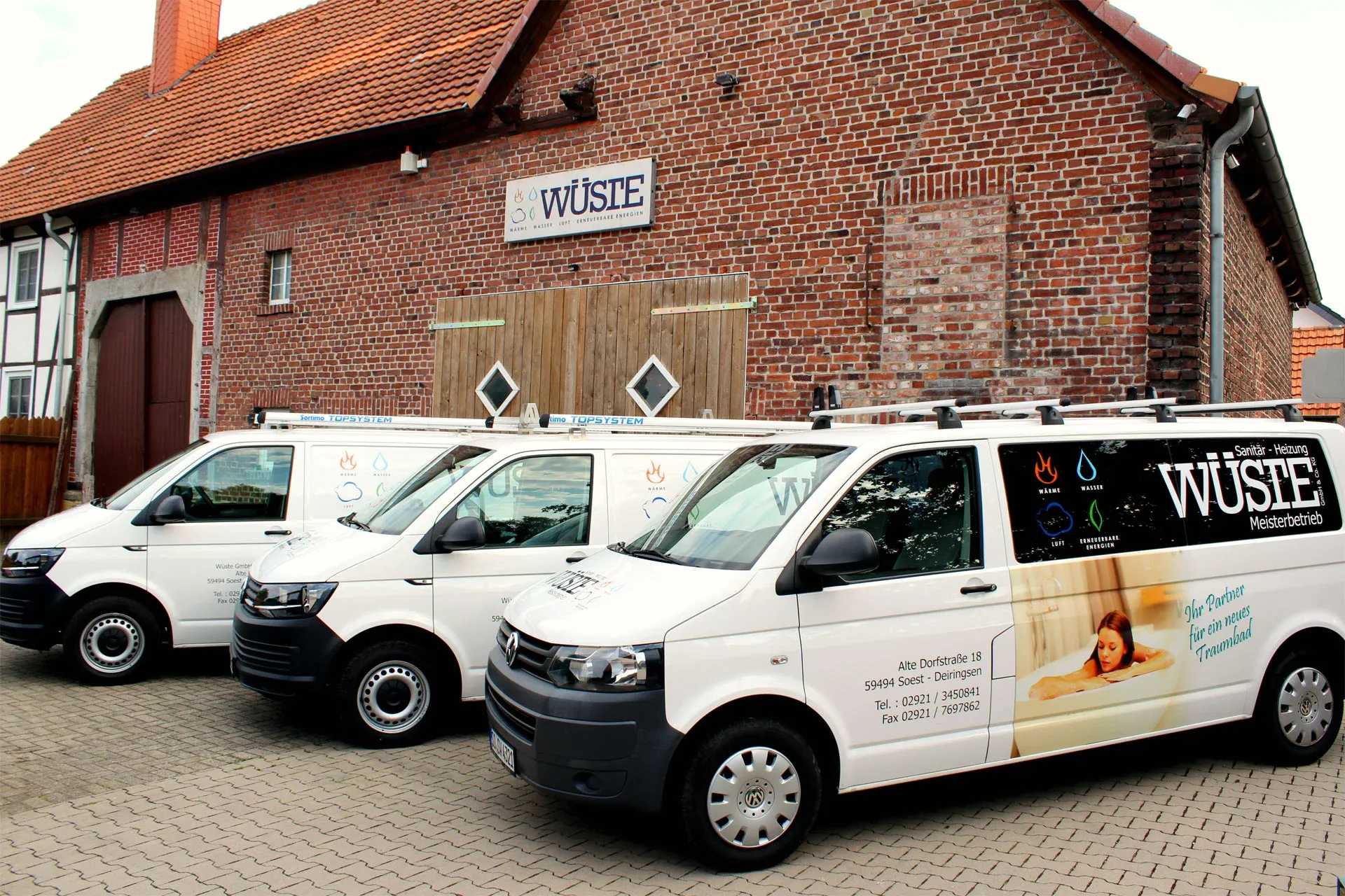 Drei weiße Servicefahrzeuge der Firma Wüste parken vor einem Backsteingebäude mit Firmenschild und rotem Ziegeldach.
